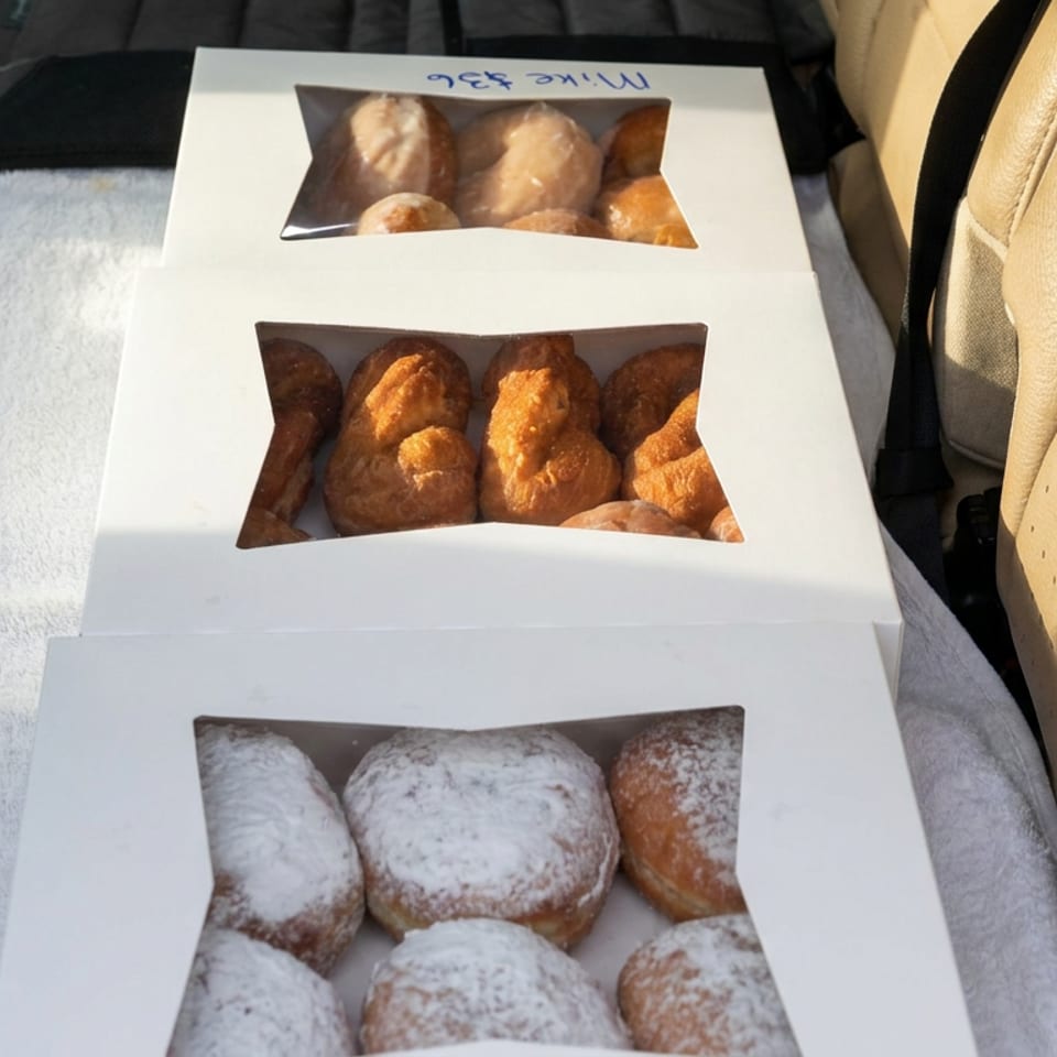 Three open pastry boxes hold assorted golden and powdered-sugar pastries on a towel inside a car.