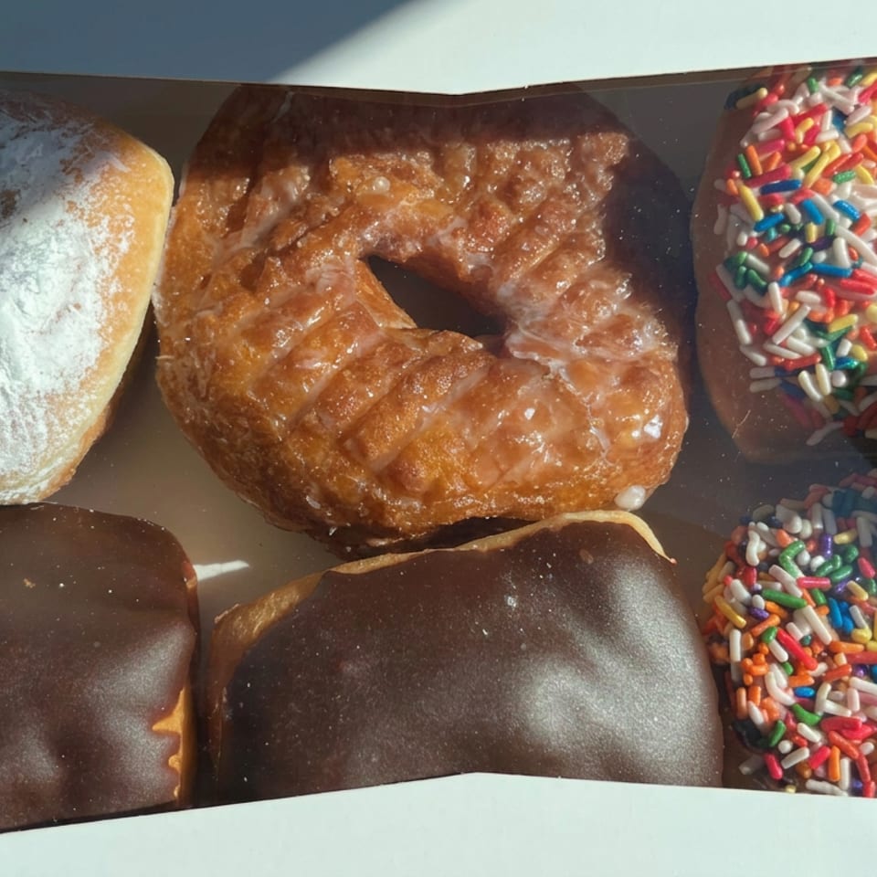 Box of assorted doughnuts, including glazed, powdered, chocolate-covered, and colorful sprinkle-topped varieties.